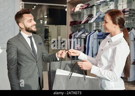 Friendly young seller of clothing store giving shopping bags to satisfied male customer Stock Photo