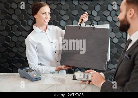 Smiling pretty store seller in blouse passing shopping bags through counter to customer while he paying cash in shop Stock Photo