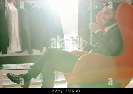 Smiling handsome young businessman in suit sitting in modern armchair in mens store and drinking coffee while using smartphone Stock Photo