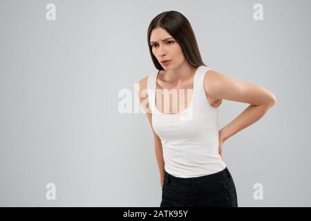 Young caucasian woman in white shirt and black jeans over gray isolated background touching back with painful expression because of ache. Front view of brunette looking down and breathing with mouth. Stock Photo