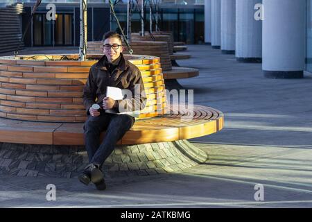 happy male Asian student programmer freelancer sitting on a bench smile drink Stock Photo