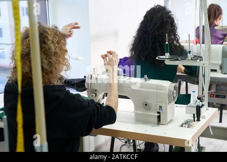Young dressmaker woman sews clothes on sewing machine. Smiling seamstress and her hand close up in workshop. Focus on sewing machine and tissue. Stock Photo