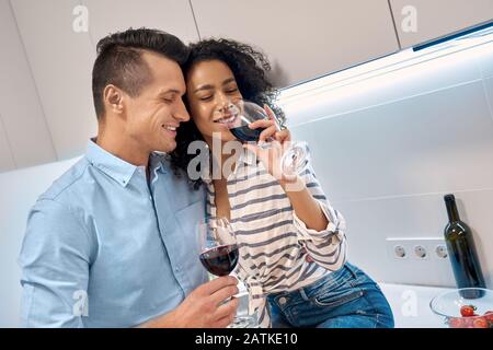 Romantic Date. Young multiethnic couple standing at kitchen cooking dinner hugging drinking wine smiling delightful Stock Photo