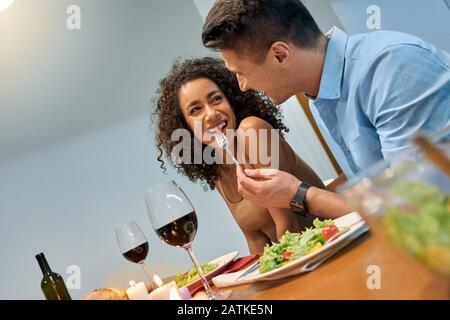 Romantic Date. Young multiethnic couple at home sitting at table eating dinner husband feeding wife with salad smiling playful Stock Photo