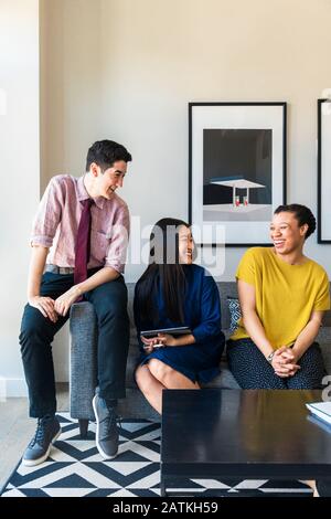 Smiling male and female business colleagues talking while sitting on sofa against wall at office Stock Photo