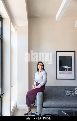 Happy female entrepreneur looking away while sitting with hands clasped on sofa against wall at office Stock Photo