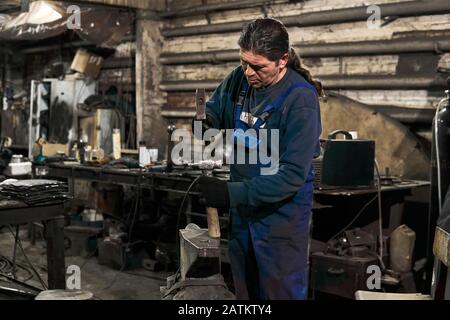 aged male blacksmith straightens a hammer in a workshop getting ready for work Stock Photo