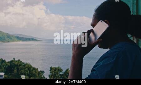 Melsisi, Pentecost Island / Vanuatu - May 10 2019: black female woman making a mobile cell phone call outside of the house in front of a beautiful sea Stock Photo