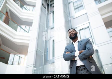 Below view of positive successful Black businessman with beard standing with crossed arms in office hall Stock Photo