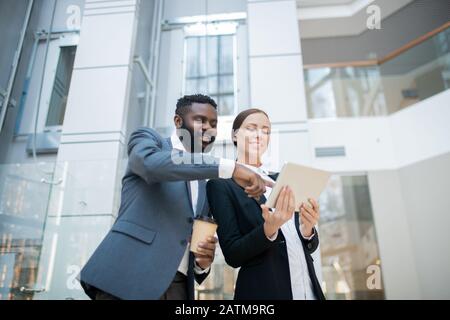 Positive young Black businessman pointing at tablet screen while discussing online project implementation with female colleague Stock Photo