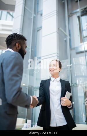 Smiling attractive young manager with tablet making handshake with Black investor while meeting him in office hall Stock Photo