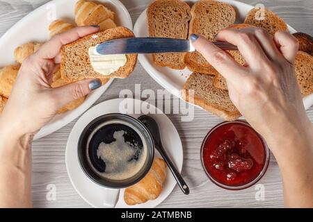 Male hands spreading butter on toast with cup of black coffee and croissant, bread, glass bowl with jam on the background. Top view. Food and beverage Stock Photo