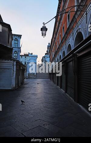 Morning on Ruga dei Oresi, roller doors down, approaching a nearly empty Rialto Bridge - Ponte di Rialto in the first light of day, Venice Italy, Stock Photo