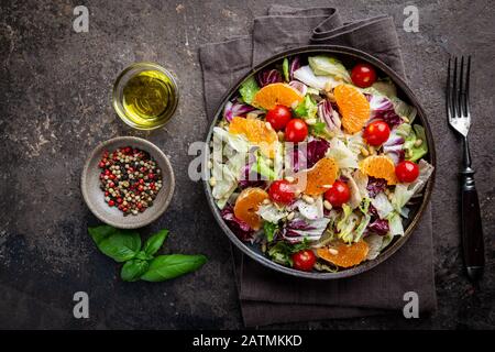 Fresh green mixed salad leaves frisee, radicchio and lamb's lettuce with tomato, olives and tangerine in a plate, top view. Stock Photo