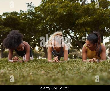 Three multiethnic smiling young active and sporty women doing plank exercises in the park - three woman being active outdoors Stock Photo