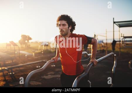 Portrait of a fitness young man listening to music on earphones doing workout in a training ground in a park - young man doing dips outdoors Stock Photo