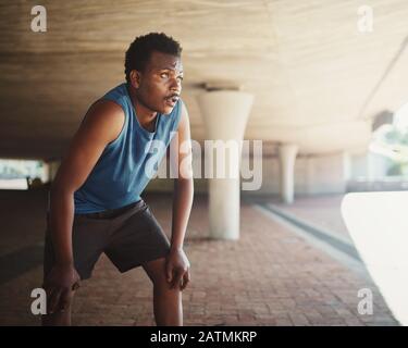 An african american male runner grasping breathe after running on street under the bridge in morning