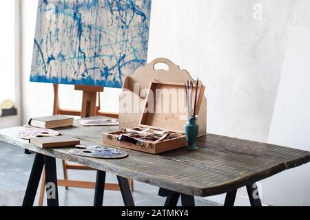 Workplace of the artist. Artwork tools on a wooden table. Clutter in artist’s workshop, watercolors, brushes, palette, easel and painting tools. Stock Photo