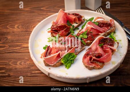 Sandwiches with prosciutto and sun-dried tomatoes on dark bread, spread with goat cheese. On a wooden background. Copy space Stock Photo