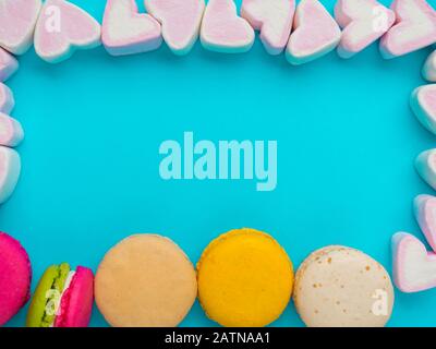 Colorful macaroons and pink marshmallow in heart shaped on trendy blue color surface, copy space. Sweet candy for love theme on Valentine concept. Top Stock Photo