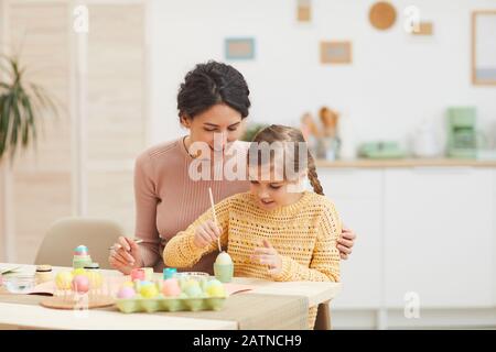 Portrait of loving mother and daughter painting Easter eggs pastel colors sitting at table in cozy kitchen interior, copy space Stock Photo