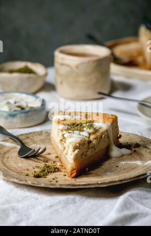 Piece of cheesecake with vanilla milk and pistachios topping on ceramic plates and variety of sauces in ceramic bowls on white table cloth. Close up. Stock Photo