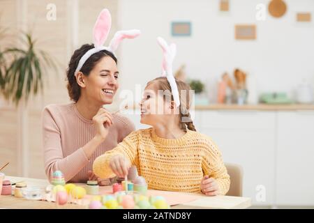 Portrait of loving mother with daughter having fun while painting Easter eggs in cozy kitchen interior, both wearing bunny ears, copy space Stock Photo