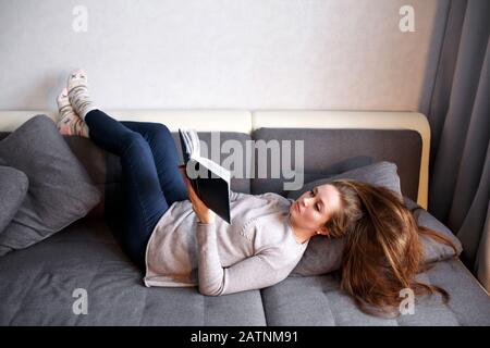 Smart young female relaxing at home, reading book on the couch. Attractive woman with long hair, minimalistic hugge interior. Stock Photo