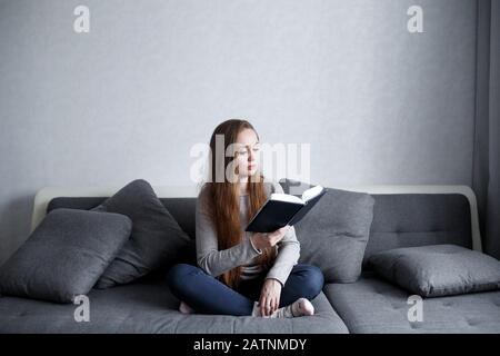 Smart young female sitting on the couch at home reading book. Attractive woman with long hair in modern minimalistic interior. Full length portrait. Stock Photo