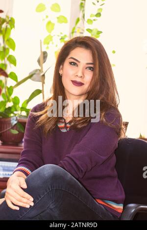 Portrait of a young pretty woman sitting on a chair in violet sweater and blue jeans with crossed legs on a sunny day with the potted plants in the ba