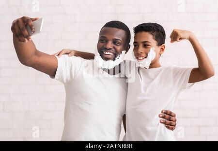 Masculine selfie. Dad and son having fun together, posing with shaving foam on cellphone camera, grey studio background Stock Photo
