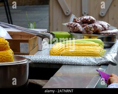 sweetcorn street vendor. stainless steel cart on street. bright yellow corn cobs in raw form. kid's hand and blurry paper money. concession stand. Stock Photo