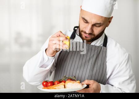 Male Chef Squeezing Lemon Plating Roasted Salmon Steak In Kitchen Stock Photo