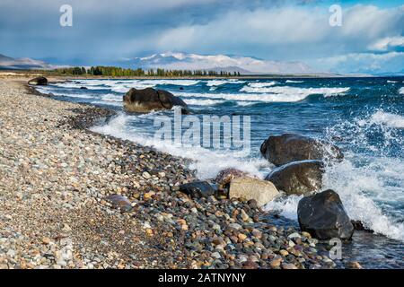 Waves breaking over rocks and pebbles at beach of Lago Buenos Aires aka Lago General Carrera, Andes mountains in far distance, Patagonia, Argentina Stock Photo