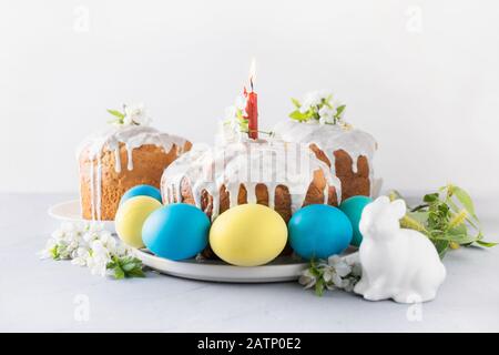 Kulich and colorful blue and yellow eggs, spring flowers on light background. Close up. Easter christianity traditional breakfast. Stock Photo