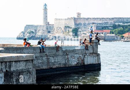 Havana, Cuba - 25 July 2018: People fishing while sitting on a cement wall located on the Malecon with El Morro Castle in the background in Havana Cub Stock Photo