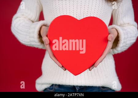 Partial view of girl in knitted sweater holding heart shaped box isolated on red background. Stock Photo