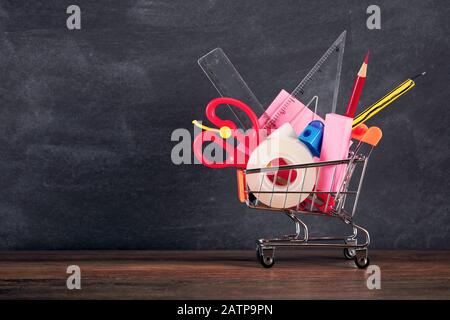 Stationery supplies in a shopping cart on a wooden desk against blackboard. Back to school concept. Stock Photo