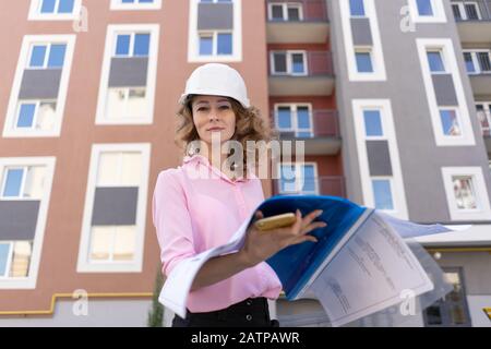 Female architect in hardhat holding folder near building. Stock Photo