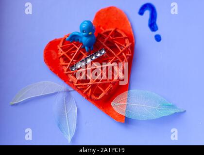 Still life, love & romance lighthearted concept -  small blue bird nested on painted red hearts with fabric feathers around edging. Copyspace. Stock Photo