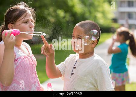 A girl stands blowing bubbles with a boy touching the bubble wand Stock Photo