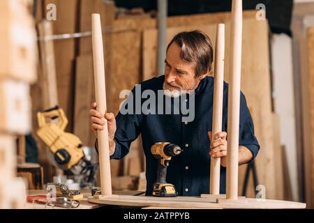 Senior man observing table legs in the workshop. Carpenter working on furniture. Stock Photo