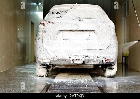 The car is covered in white foam under a stream of spray from the bottom to the bottom in a car wash, rear view. Stock Photo