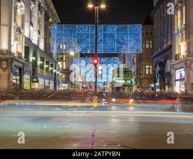 Christmas lights on Oxford Street at night with a green bus stopped at traffic lights with cars and people moving around. Long exposure photo. London Stock Photo