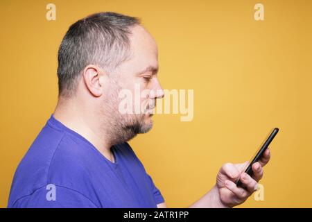 side view of mature man using and looking at smartphone or mobile smart phone reading text message - yellow studio background with copy space Stock Photo