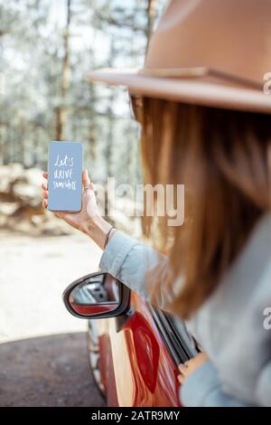 Woman traveling by car in the forest, leaning out of the car window holding smartphone with text calling to travel Stock Photo