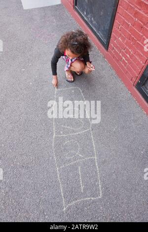 A girl crouches on the sidewalk and draws hopscotch game with chalk Stock Photo