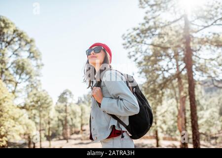 Lifestyle portrait of a stylish woman dressed casually in red hat walking with backpack in the forest