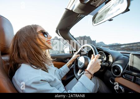 Happy woman driving convertible car while traveling on the desert road. Carefree lifestyle and travel concept