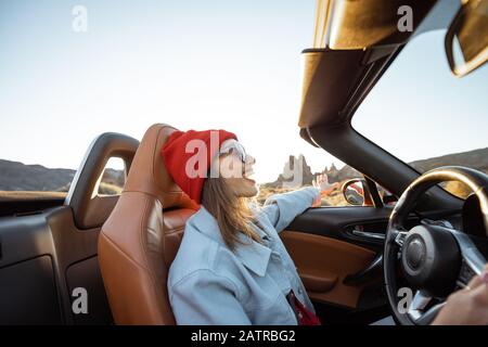 Happy woman in red hat driving convertible car while traveling on the desert road with beautiful rocky landscape on the background during a sunset